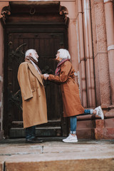 Side view full length portrait of old bearded gentleman and his wife standing near door of old building. They looking at each other and smiling