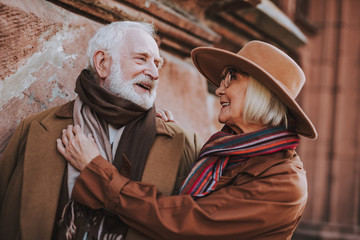 Portrait of happy elderly lady in hat hugging husband while looking at him and smiling