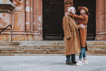 Side view full length portrait of stylish bearded man embracing his wife while standing near old building. They looking at each other and smiling - obrazy, fototapety, plakaty