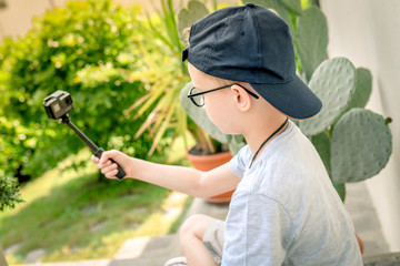 Young boy taking selfie on action camera outdoor with a cactus in background. Caucasian pre teen playing with action cam in the garden. Beautiful kid with hat and glasses.