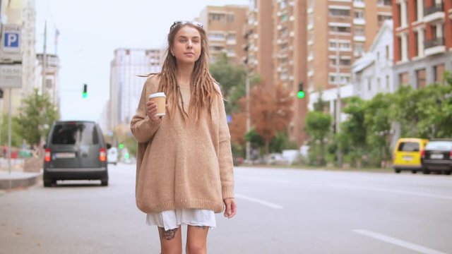 young caucasian woman walking along the road in urban city slow motion. caucasian model with dreadlocks wearing oversized sweater holding cup of coffee drinking on the run outdoors