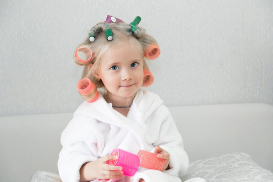 Girl with curlers hairpins in her hair. A little girl in a white coat spun pink curlers into her hair.