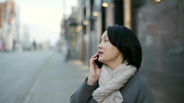 Tilt down medium shot of stylish middle-aged Asian woman having phone conversation while standing outdoors