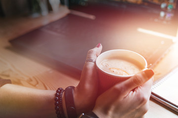 Student woman holds with two hands a hot cup of tea with sea buckthorn Hippophae and a slice of orange. Workplace, notebook with pen and laptop, the working process, warm color toning, space for text