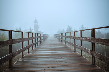 Fototapeta premium Fabulous blue mist. Empty wooden bridge stretching into the mystical fog. A watchtower looms in the background in the distance. Cold autumn morning.