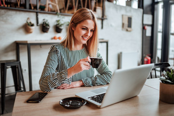Young female entrepreneur drinking cup of coffee and looking at laptop screen.