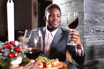Portrait of African American businessman with a glass of wine in his hand. Businessman in a suit and in the interior of the restaurant with Christmas accessories on the background of the fireplace.