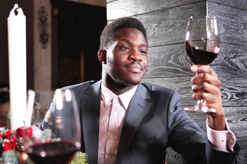 Portrait of African American businessman with a glass of wine in his hand. Businessman in a suit and in the interior of the restaurant with Christmas accessories on the background of the fireplace.