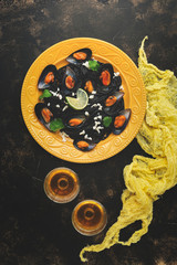 Black pasta spaghetti with seafood mussels on a yellow plate with white wine, textiles, dark rustic background. Top view, flat lay.