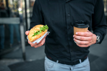 Cut view of man holding salty croissan and cup of drink. He stands at building. Guy wears black shirt and blue jeans.
