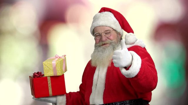 Cheerful Santa giving thumb up. Old Santa Claus with real beard holding gift boxes on blurred background. Special Christmas offer.