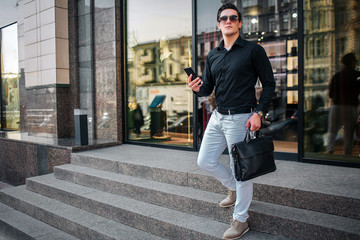 Young businessman pose on camera. He walks down on steps. Guy holds phone and black bag. Young man is outside.