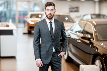 Portrait of a handsome salesman in the suit standing at the showroom with luxury cars on the background