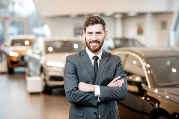 Portrait of a handsome salesman in the suit standing at the showroom with luxury cars on the...