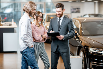 Obraz premium Handsome male dealer selling to a young couple clients a new car, standing together with some documents in the showroom