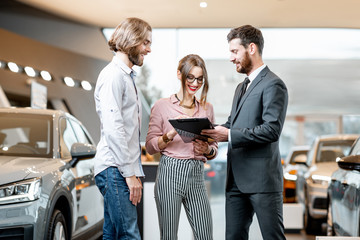 Handsome male dealer selling to a young couple clients a new car, standing together with some...