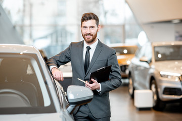 Portrait of a handsome salesman in the suit standing near the car in the showroom
