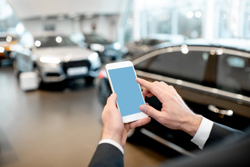 Businessman holding phone with empty screen to copy space in the showroom with cars on the background