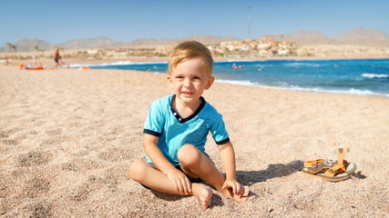 Portrait of cute three years old toddler boy sitting on the sandy beach at sea.