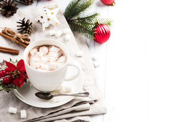 Cup of hot chocolate with marshmallow and cinnamon sticks on the white wooden table. Winter cocoa drink on a napkin, Christmas-tree golden stars and red balls decorations, branches and cones