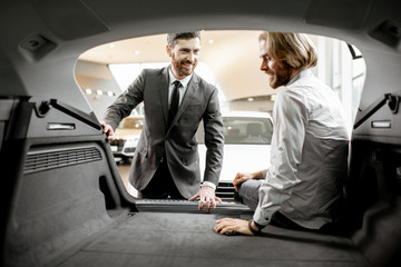 Salesman showing spacious trunk of a new car for a young man client in the showroom