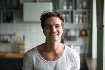 Fototapeta premium Head shot portrait of handsome confident cheerful male in casual clothes at home opposite kitchen looking at camera. Young millennial positive man work at home, freelance or have free time on weekend