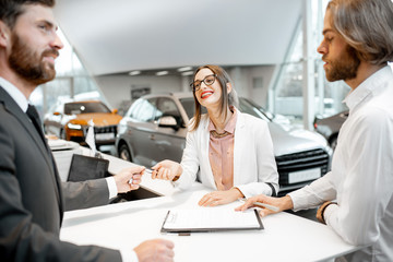 Young business couple signing some documents at the table with salesperson or manager buying or renting car in the showroom