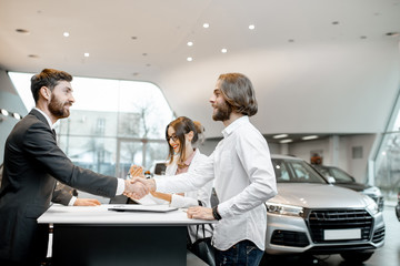 Young business couple shaking hands having a deal with salesperson at the stand buying or renting car in the showroom