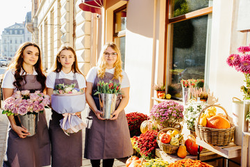 Three friendly florists female team holding different flowers in pots outdoor near shop window with autumn decoration of their floral shop. Small business concept.