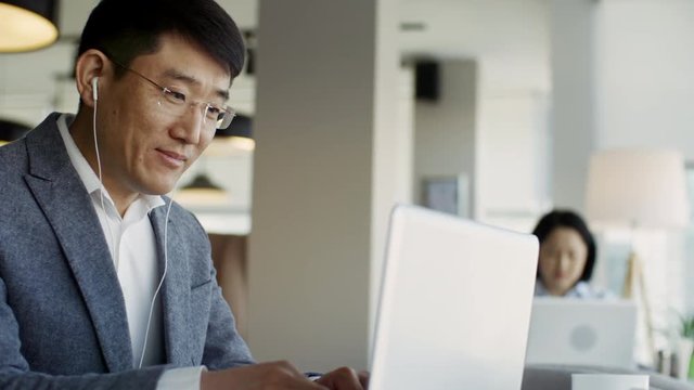 Tracking medium shot of middle-aged Asian man in glasses using laptop computer and earphones during video conversation with colleague