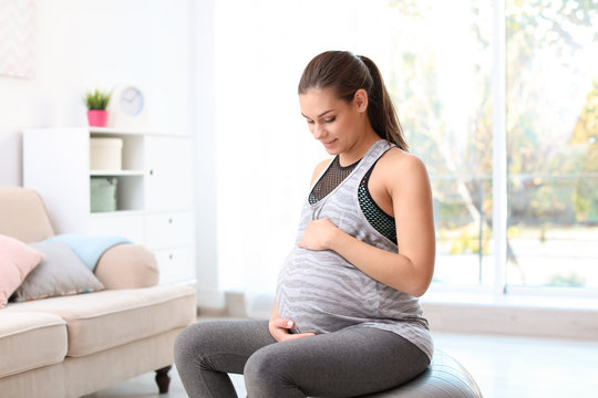 Young pregnant woman in fitness clothes sitting on exercise ball at home