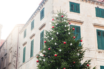 Fototapeta na wymiar Christmas tree with red ornament, white snowflakes outside in sun light. Street town decoration near stone building with windows, wooden shutters. Authentic festive atmosphere in Dubrovnik Croatia