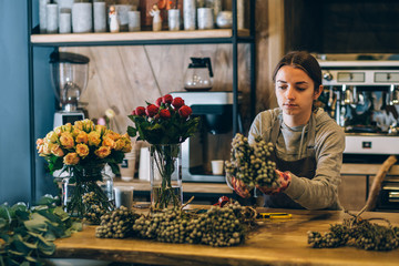 Atmosphere photo of smiling brunette lovely female business woman florist standing behind wooden counter and creating beautiful bouquet wit burgundy roses in modern loft interior floral shop.