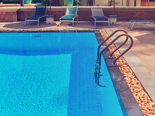 Sun beds are near the pool without people. The pool is filled with clean water. Metal ladder is on...