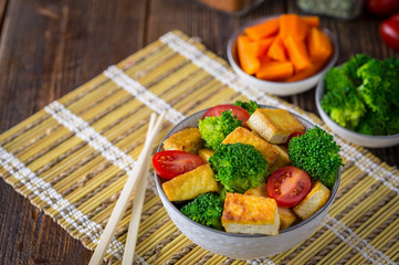 Grilled tofu with broccoli and tomatoes in white bowl on bamboo pad with chopsticks on the left side.