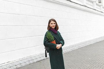 Beautiful young stylish brunette woman, wearing green coat, walking through the city streets with small Christmas tree in her hands. New year concept. Christmas tree in a pot.