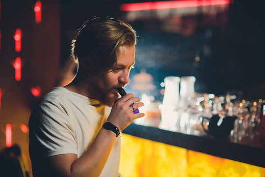 Young handsome hipster man with beard sitting in cafe with a cup of coffee, vaping and releases a cloud of vapor. - Powered by Adobe