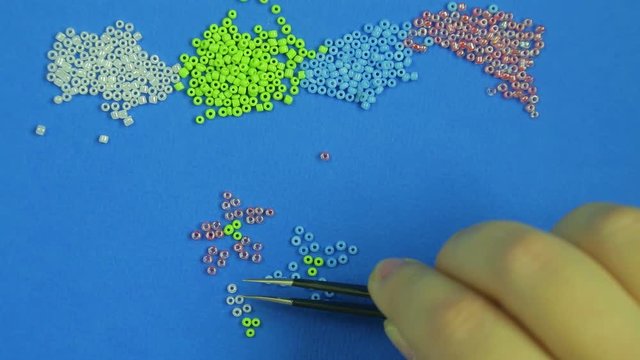 Female hand on a blue background pokes out flowers from multi-colored beads with tweezers. Close-up. Handmade