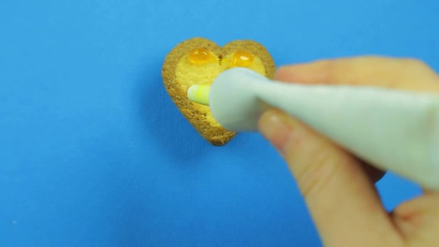 Female hand paints heart-shaped cookies on a blue background with yellow icing from a tube funny faces.