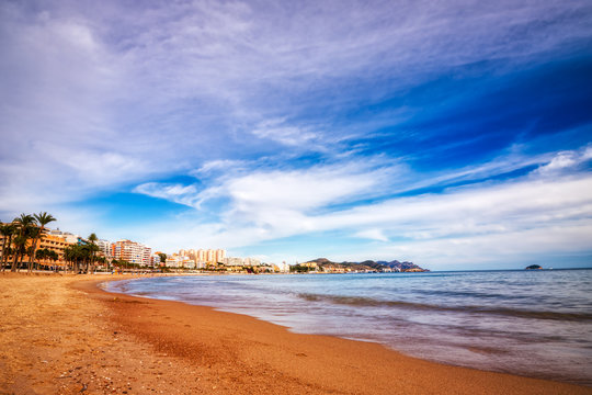 Low angle shot of the waves coming up on the snady beach of Villajoyosa Spain with a blue sky, white clouds and the buildings lining the shore in the distance.