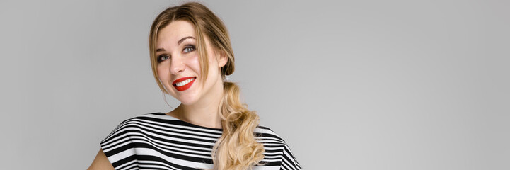 Naklejka premium Attractive young blonde girl in striped blouse smiling holding her hair standing on gray background