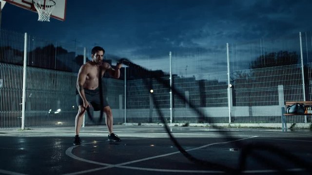 Strong Muscular Fit Shirtless Young Man is Doing Exercises with Battle Ropes. He is Doing a Workout in a Fenced Outdoor Basketball Court. Evening Footage After Rain in a Residential Neighborhood Area.