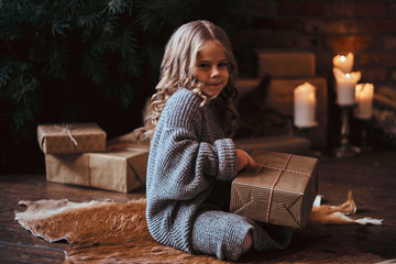 Cute little girl with blonde curly hair wearing a warm sweater holding a gift box while sitting on...