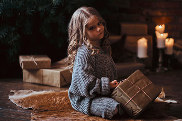 Dissatisfied little girl with blonde curly hair wearing a warm sweater sitting on a floor...