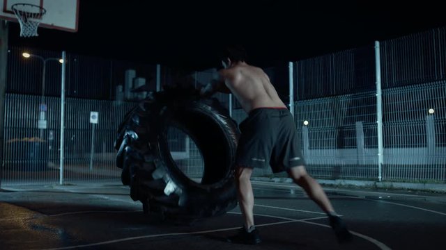 Backshot of a Strong Muscular Fit Young Shirtless Man Doing Exercises in a Fenced Outdoor Basketball Court. He's Flipping a Big Heavy Tire in a Night After Rain in a Residential Neighborhood Area.