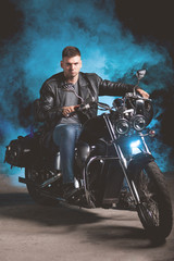 Fototapeta na wymiar Cute biker in leather jacket sits on a motorcycle in blue smoke