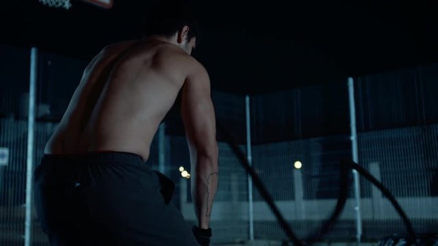 Backshot of a Strong Muscular Fit Shirtless Young Man Doing Exercises with Battle Ropes. He is Doing a Workout in a Fenced Outdoor Basketball Court. Night Footage After Rain in a Neighborhood Area.