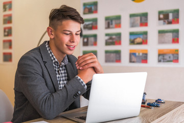 Portrait of the smiling young man who is sitting at the table and working with laptop indoor