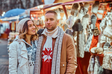 Fototapeta premium A young romantic couple walking on the street at Christmas time, standing near decorated shops in the evening, enjoying spending time together.