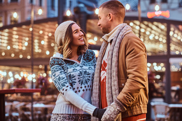 A young romantic couple holding hands and looking at each other while standing on the street at Christmas time, enjoying spending time together.